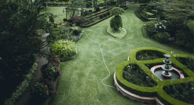 A beautifully landscaped garden featuring a neatly trimmed lawn, meticulously shaped hedges, and a central fountain surrounded by a circular hedge design. The garden also showcases diverse plant life and structured pathways, reflecting the professional work of Pines Landscaping. The overall design emphasizes both aesthetic appeal and functional use of space, providing a serene and visually pleasing environment.