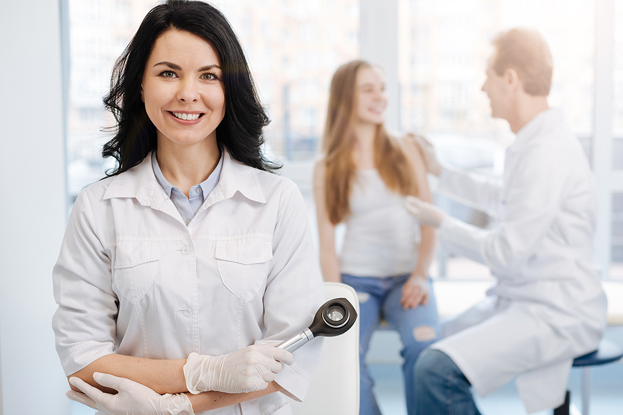 With individual attitude to every patient. Smiling charming beautiful dermatologist working in the clinic and holding dermatoscope while colleague talking to young patient in the background